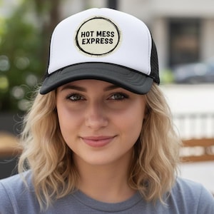 Könnte beinhalten: Eine weiß-schwarze Trucker-Cap mit einem runden Aufnäher auf der Vorderseite, der einen abgenutzten Rand aufweist. Der Aufnäher zeigt den schwarzen Schriftzug HOT MESS EXPRESS in einer fetten, serifenlosen Schrift auf cremefarbenem Grund.