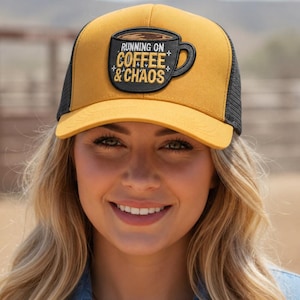 Puede incluir: Una gorra tipo camionero de color amarillo mostaza y negro con un parche negro en forma de taza de café. El parche muestra el texto RUNNING ON COFFEE & CHAOS en letras amarillas y blancas, decorado con pequeñas estrellas sobre un fondo oscuro.