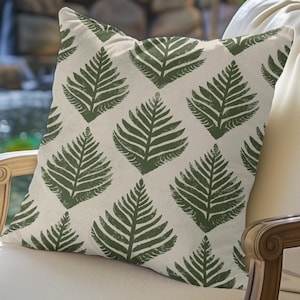 May include: Square decorative pillow with a repeating pattern of dark green fern leaves on a cream background. The pillow is placed on a white chair with a wooden frame, suggesting outdoor or garden decor.