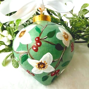 May include: A teardrop-shaped green Christmas bauble featuring hand-painted white dogwood flowers with yellow centres and small red berries on brown branches. The bauble has a gold-coloured metal cap and a white satin ribbon bow.