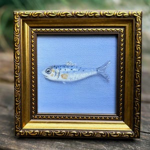 May include: A small, square, ornate gold-colored frame containing a painting of a single fish. The fish is rendered in shades of blue, silver, and white with a small gold spot on its side, positioned horizontally against a solid light blue background.
