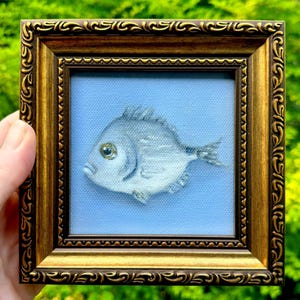 May include: A small square painting of a silver fish centered on a light blue background, enclosed in an ornate, dark gold-toned frame with a decorative embossed border pattern. The fish is depicted in profile with a single visible eye.