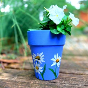 May include: A bright blue terracotta flower pot featuring hand-painted white daisy flowers with yellow centers and green stems. The pot contains a small green plant with white five-petaled flowers blooming from the top of the container.