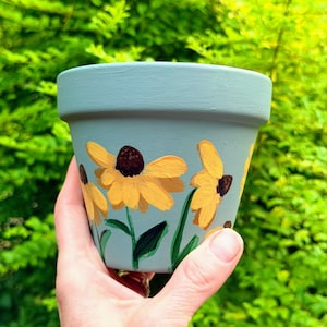 May include: A light blue flower pot featuring hand-painted yellow coneflowers with dark brown centers and green stems. The pot has a wide, flat rim and a smooth, matte finish, showcasing a floral design wrapped around the exterior surface.