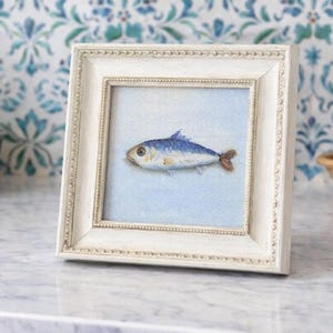 May include: A square, off-white wooden frame with a beaded inner edge containing a small canvas painting of a single fish. The fish is rendered in shades of blue, silver, and brown, set against a solid, light blue background within the frame.
