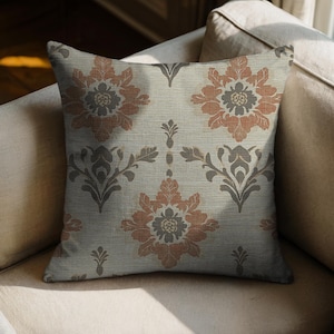 May include: Square decorative pillow with a floral pattern in shades of rust, gray, and cream. The pillow features a repeating design of stylized flowers and leaves, creating a classic and elegant look. The pillow is placed on a beige sofa.