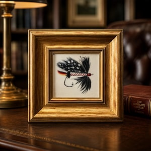May include: A framed artwork featuring a detailed fly fishing lure. The lure has black and white speckled wings, red accents, and a curved hook. The artwork is set in a gold-colored, square frame, and is displayed on a wooden surface.