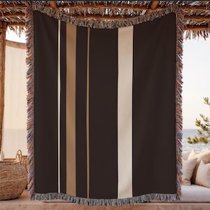 May include: A dark brown throw blanket with vertical stripes in beige and tan. The blanket has a fringed edge with multiple colors. The blanket is hanging outdoors, possibly on a patio or deck.