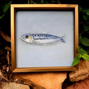 May include: A square framed painting featuring a single fish centered on a light blue canvas. The fish is rendered in shades of blue, silver, and white with visible brushstrokes, set within a metallic gold frame that surrounds the artwork.