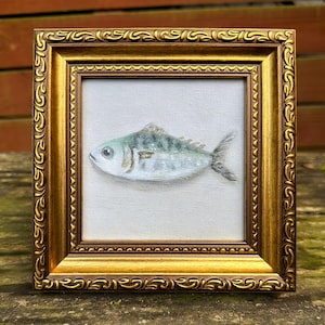 May include: A small square painting of a fish with silver and green scales, centered on a white canvas. The artwork is enclosed in an ornate, metallic gold-toned frame featuring a decorative embossed scroll pattern along the outer border.