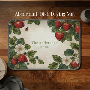 Peut inclure: Tapis de séchage de la vaisselle absorbant rectangulaire avec un motif de fraises et de fleurs. Le tapis a un fond crème avec des fraises rouges, des feuilles vertes et des fleurs blanches. Le texte "The Andersons EST 2014" est imprimé sur le tapis. Le tapis mesure 53 cm.