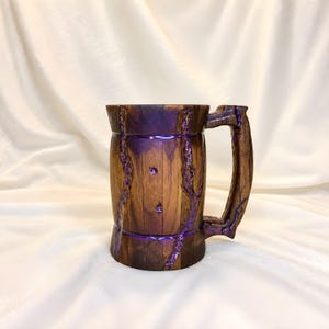 May include: A wooden beer mug featuring a barrel-like design with vertical staves and a sturdy handle. The mug is accented with deep purple resin inlays that fill the crevices and seams, creating a smooth, glossy contrast against the brown wood.