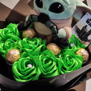 Op de afbeelding: Een uniek boeket met een Grogu-knuffel omringd door groene satijnen rozen. Goudfolie verpakte chocolaatjes en sprankelende details verfraaien het arrangement, verpakt in zwart papier. Een Star Wars-label is zichtbaar.