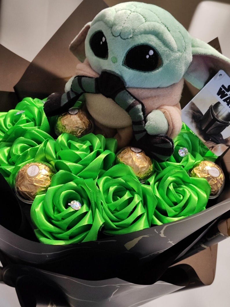 Op de afbeelding: Een uniek boeket met een Grogu-knuffel omringd door groene satijnen rozen. Goudfolie verpakte chocolaatjes en sprankelende details verfraaien het arrangement, verpakt in zwart papier. Een Star Wars-label is zichtbaar.