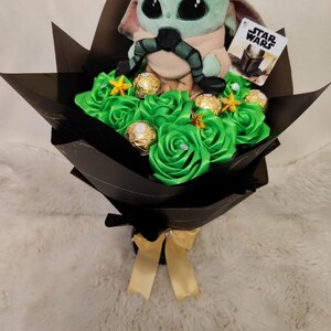 Op de afbeelding: Een boeket met een Grogu-knuffel uit Star Wars, groene satijnen rozen, goud omwikkelde chocolade en stervormige versieringen. Het boeket is verpakt in zwart papier en vastgebonden met een gouden lint. Een Star Wars-kaart is bevestigd.
