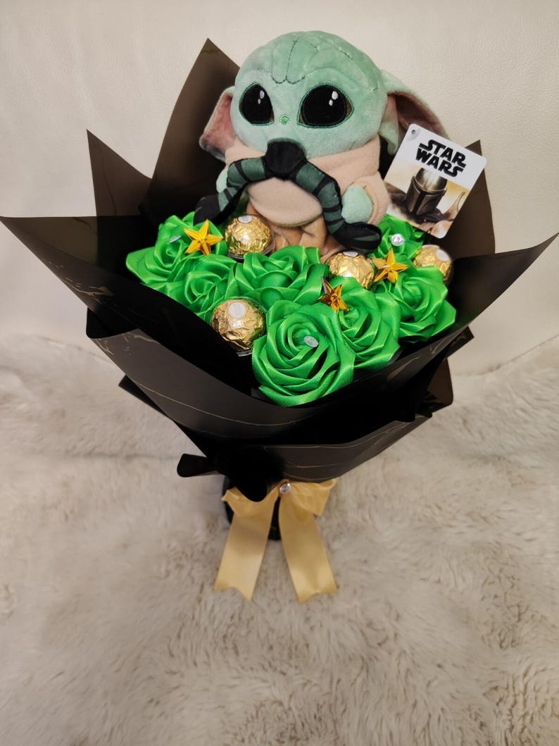 Op de afbeelding: Een boeket met een Grogu-knuffel uit Star Wars, groene satijnen rozen, goud omwikkelde chocolade en stervormige versieringen. Het boeket is verpakt in zwart papier en vastgebonden met een gouden lint. Een Star Wars-kaart is bevestigd.