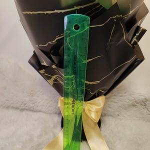 Op de afbeelding: Een boeket verpakt in zwart papier met gouden aderen, vastgebonden met een gouden lint. Een groene liniaal toont dat het boeket ongeveer 25 cm hoog is. Het boeket bevat een knuffel.