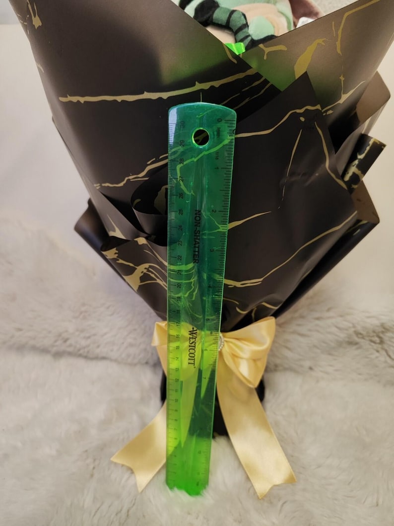 Op de afbeelding: Een boeket verpakt in zwart papier met gouden aderen, vastgebonden met een gouden lint. Een groene liniaal toont dat het boeket ongeveer 25 cm hoog is. Het boeket bevat een knuffel.