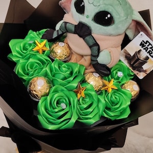 Op de afbeelding: Een boeket met een Grogu-knuffel, groene rozen van lint, goud verpakte chocolaatjes en stervormige versieringen. Het boeket is verpakt in zwart papier en vastgebonden met een gouden lint.