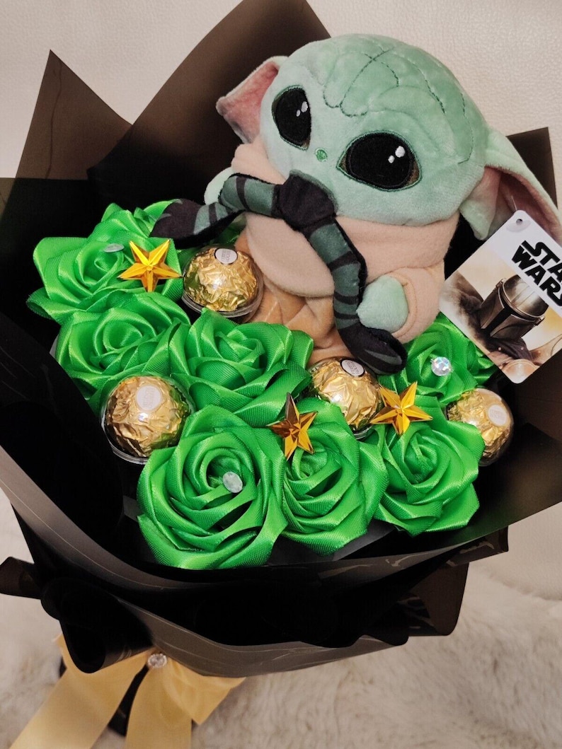 Op de afbeelding: Een boeket met een Grogu-knuffel, groene rozen van lint, goud verpakte chocolaatjes en stervormige versieringen. Het boeket is verpakt in zwart papier en vastgebonden met een gouden lint.