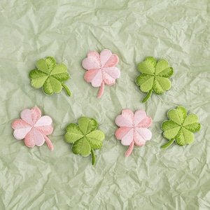 Pode incluir: Remendos de trevo bordados em tons de verde e rosa, dispostos sobre uma superfície texturizada verde claro. Os remendos apresentam costuras detalhadas e um caule. A imagem mostra uma coleção de apliques decorativos.