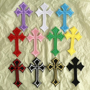 May include: Eleven embroidered cross patches in a variety of colours, including purple, white, blue, red, pink, green, yellow, black, and black with white and yellow trim. Each cross features a detailed design and a contrasting border.