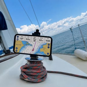 Może przedstawiać: Smartfon zamontowany na kabestanie żaglówki, wyświetlający aplikację nawigacyjną z danymi takimi jak 127 stopni COG, 6,4 węzła SOG, 14 węzłów wiatru SW, 8,2 metra głębokości i 14:35 ETA. Kabestan jest owinięty wielokolorową liną.