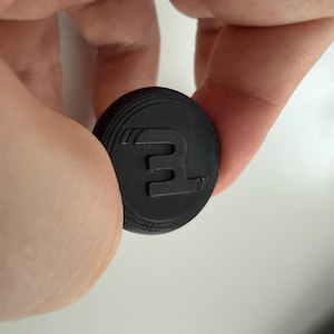 Puede incluir: Un objeto negro y circular con un diseño en relieve que presenta la letra "E" y una forma de ala. El objeto tiene un borde estriado y se sostiene entre los dedos.