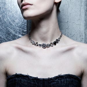 Puede incluir: Un collar plateado con varios elementos decorativos, incluyendo piedras negras y diseños circulares. El collar se usa sobre un top de mezclilla y una pared gris texturizada.
