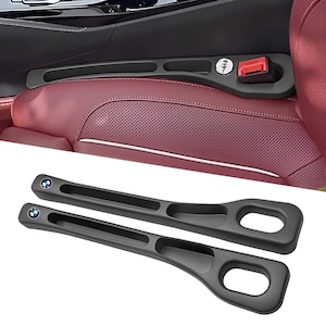 May include: Two black car seat belt extenders with the BMW logo, designed to fit into a car's interior. The extenders have a sleek, modern design with a textured surface and a loop for easy handling. The background shows a car's interior with a red seat.