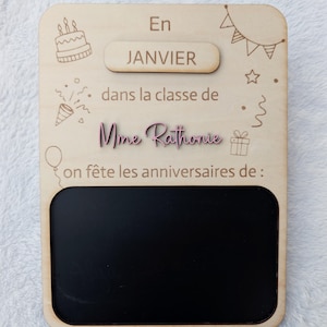 Peut inclure: Une plaque d'anniversaire rectangulaire en bois clair avec le texte gravé "En JANVIER dans la classe de Mme Rathonie on fête les anniversaires de :", des illustrations de gâteau, guirlandes, étoiles, cadeau et une zone tableau noir.