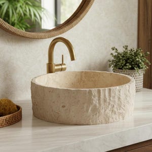 Op de afbeelding: Een beige stenen wastafel met een getextureerde buitenkant staat op een wit aanrechtblad. Een gouden kraan is boven de wastafel gemonteerd. Een ronde spiegel met een geweven frame hangt aan de muur. Een kleine plant en decoratieve items staan op het aanrecht.