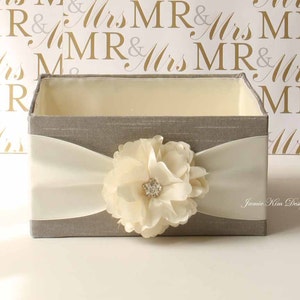 May include: A gray fabric box with a white satin ribbon and a large ivory flower with a rhinestone center. The box is on a white surface with a background of a repeating pattern of "MR & Mrs" in gold and gray.