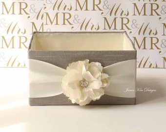 May include: A gray fabric box with a white satin ribbon and a large ivory flower with a rhinestone center. The box is on a white surface with a background of a repeating pattern of "MR & Mrs" in gold and gray.