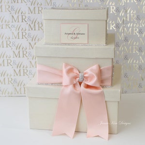 May include: Three white gift boxes with a pink satin bow and rhinestone trim. The top box has a personalized label with the names "Arianna & Adriano" and the date "May 24, 2014".