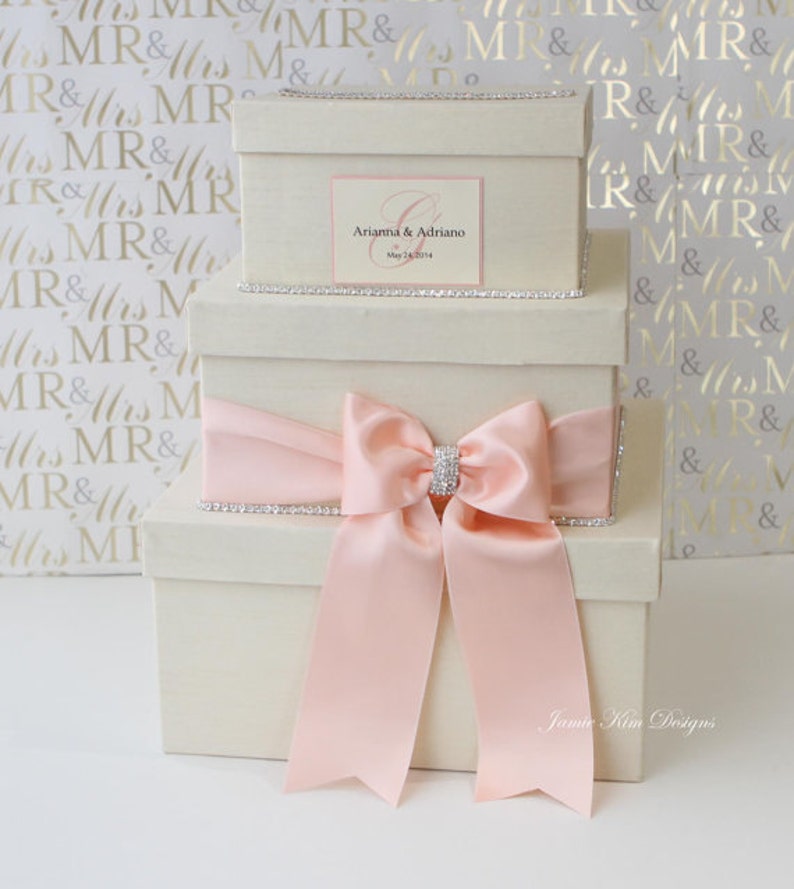 May include: Three white gift boxes with a pink satin bow and rhinestone trim. The top box has a personalized label with the names "Arianna & Adriano" and the date "May 24, 2014".