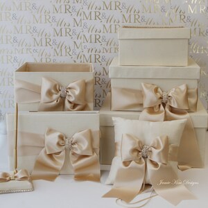 May include: Set of five ivory boxes with satin ribbon bows and rhinestone accents. The boxes are arranged in a pyramid shape, with the largest box at the bottom and the smallest box at the top. The boxes are perfect for storing wedding cards, gifts, or other special items.