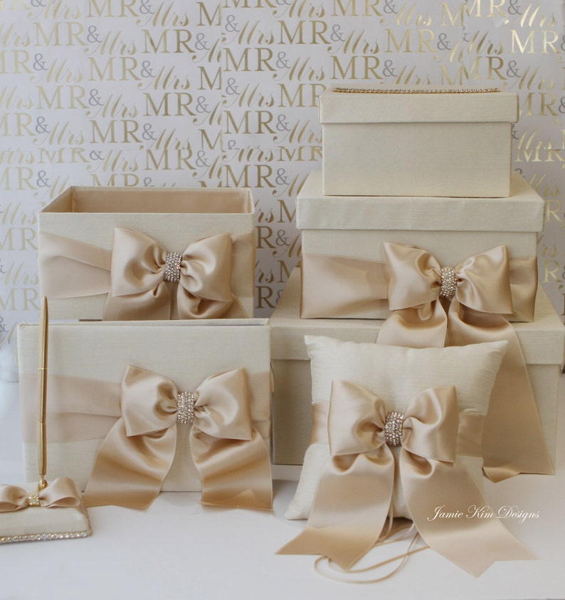 May include: Set of five ivory boxes with satin ribbon bows and rhinestone accents. The boxes are arranged in a pyramid shape, with the largest box at the bottom and the smallest box at the top. The boxes are perfect for storing wedding cards, gifts, or other special items.