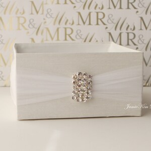 May include: A white square box with a white tulle ribbon and a rhinestone embellishment. The box is sitting on a white surface with a gold background.