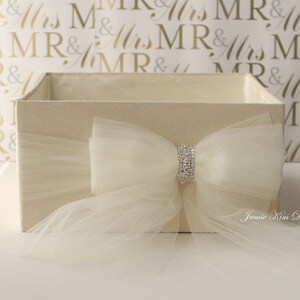 May include: A white gift box with a large ivory tulle bow and a silver rhinestone embellishment. The box is sitting on a white surface with a gold and white background.