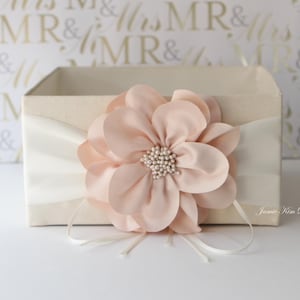 May include: A white box with a blush pink flower on top. The box has a white satin ribbon around it. The text "Jamie Kim Designs" is printed on the box.