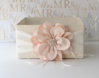 May include: A white box with a blush pink flower on top. The box has a white satin ribbon around it. The text "Jamie Kim Designs" is printed on the box.