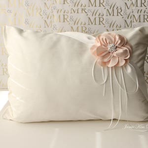 Könnte beinhalten: Ein weißes Kissen mit einer pfirsichfarbenen Blume und einem weißen Satinband. Das Kissen liegt auf einer weißen Oberfläche mit goldenem Hintergrund.