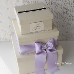 May include: A three-tiered wedding card box with a light beige finish. The top tier has a slot for cards and is decorated with rhinestones. The middle tier has a personalized label with the names "Luke & Stacey" and the date "15 May 2014". The bottom tier has a large lavender satin bow with a rhinestone embellishment. The box is labeled "Jamie Kim Designs".