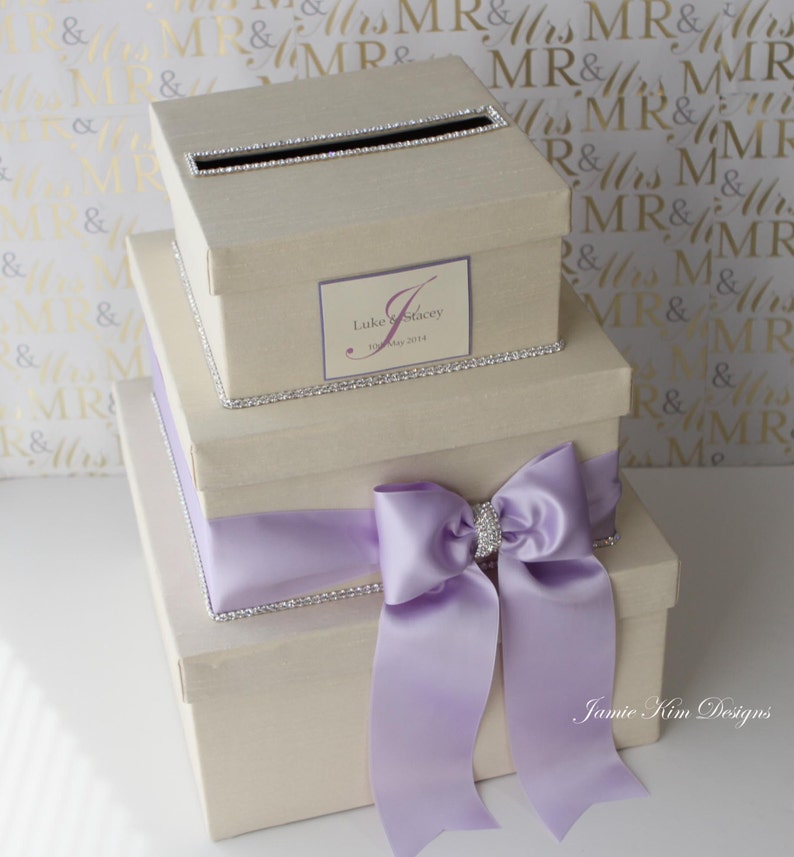 May include: A three-tiered wedding card box with a light beige finish. The top tier has a slot for cards and is decorated with rhinestones. The middle tier has a personalized label with the names "Luke & Stacey" and the date "15 May 2014". The bottom tier has a large lavender satin bow with a rhinestone embellishment. The box is labeled "Jamie Kim Designs".