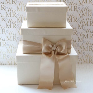 Puede incluir: Tres cajas de regalo de color marfil con una cinta de sat&eacute;n y un lazo de strass. Las cajas est&aacute;n apiladas una encima de la otra. El fondo es un patr&oacute;n blanco con las palabras "MR & Mrs" repetidas.
