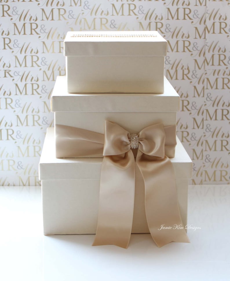 Puede incluir: Tres cajas de regalo de color marfil con una cinta de sat&eacute;n y un lazo de strass. Las cajas est&aacute;n apiladas una encima de la otra. El fondo es un patr&oacute;n blanco con las palabras "MR & Mrs" repetidas.
