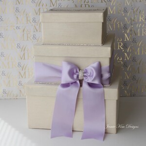 Puede incluir: Tres cajas de regalo de color marfil apiladas una encima de la otra. La caja superior es m&aacute;s peque&ntilde;a que las dos cajas inferiores. Las cajas est&aacute;n decoradas con un borde plateado y una cinta de sat&eacute;n lavanda con un lazo de pedrer&iacute;a.