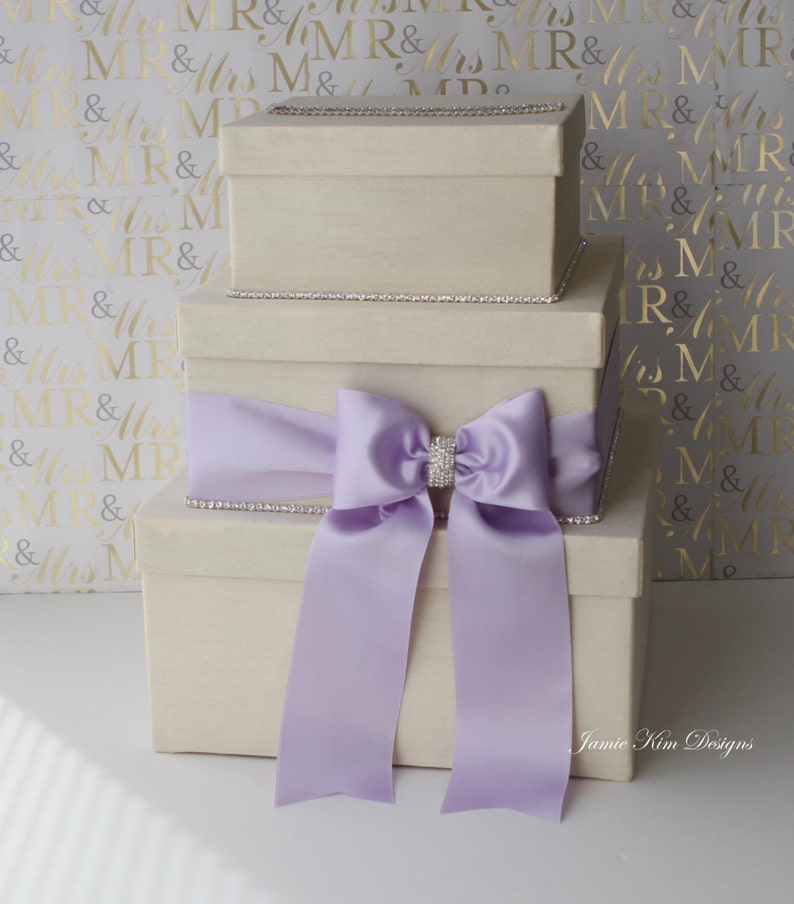 Puede incluir: Tres cajas de regalo de color marfil apiladas una encima de la otra. La caja superior es m&aacute;s peque&ntilde;a que las dos cajas inferiores. Las cajas est&aacute;n decoradas con un borde plateado y una cinta de sat&eacute;n lavanda con un lazo de pedrer&iacute;a.