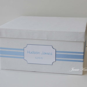 May include: White keepsake box with blue stripes and a label that reads "Hudson James 5.09.15". The box is decorated with a blue ribbon and the words "Jamie Kim Designs" are printed on the bottom right corner.